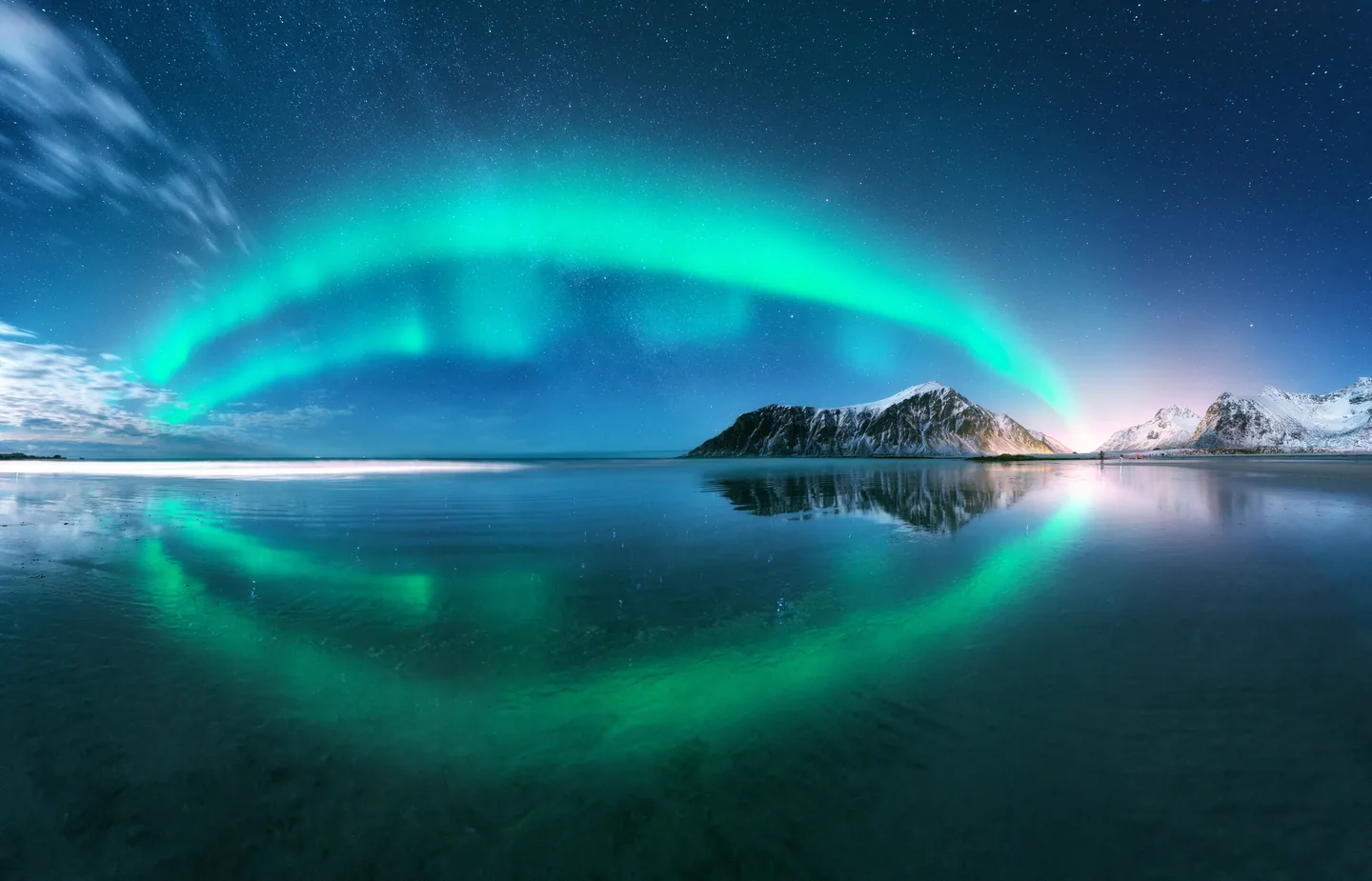 Green aurora forms an arch above mountains and reflective water in Norway.