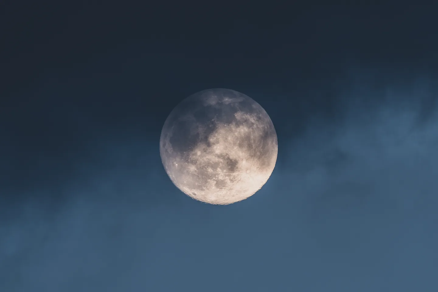 A bright Moon surrounded by thin clouds in a blue-grey night sky.